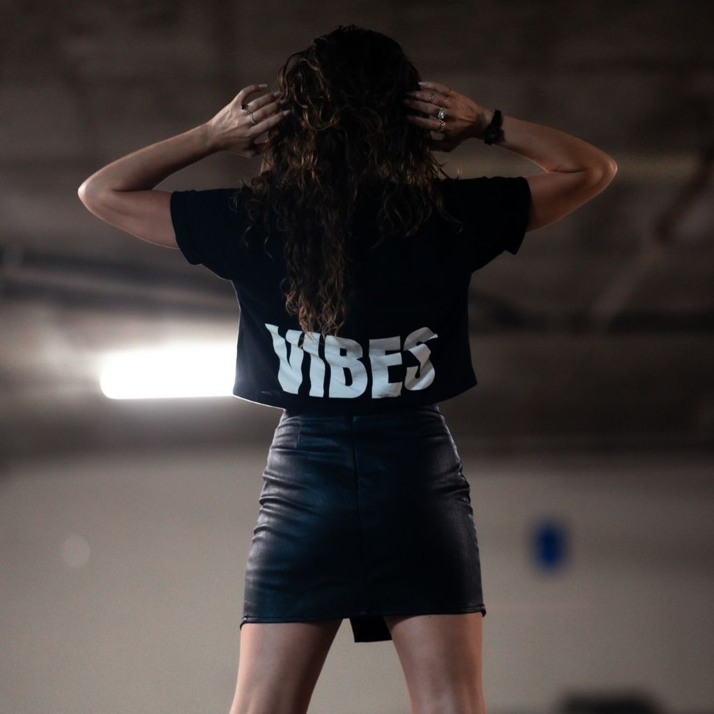 Image features the back of a female model wearing a black leather mini skirt and the black Be Kind Vibes Be Kind crop top. The model is standing in a parking garage.