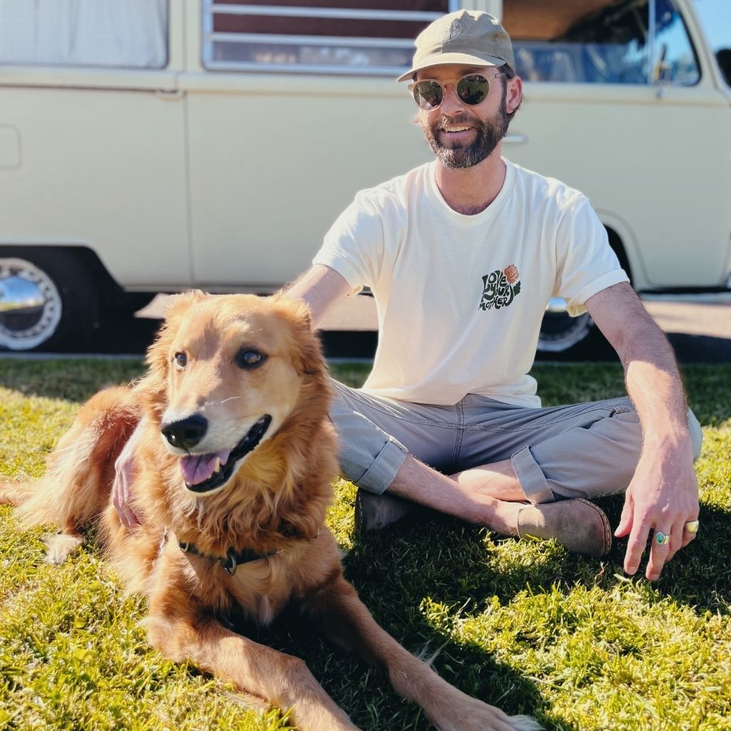 Image features the sustainably made 100% organic cotton Love Your Mother tee from Be Kind Vibes on a male model wearing sunglasses, a green hat, khaki pants and sitting in the grass with a cute golden retriever.