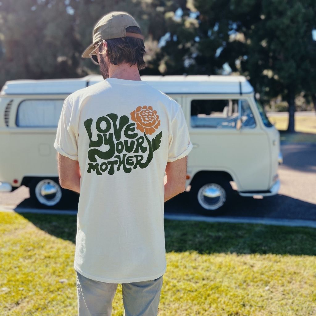 Hero image featuring the back of the 100% organic cotton Be Kind Vibes Love Your Mother tee in natural on a male model standing in front of a VW bus.