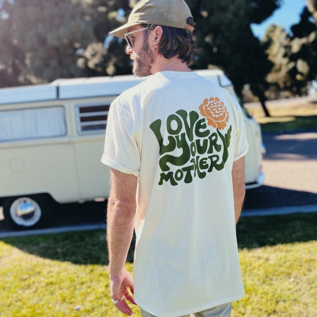 Hero image featuring the back of the 100% organic cotton Be Kind Vibes Love Your Mother tee in natural on a male model standing in front of a VW bus.