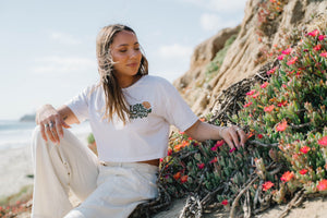 Hero image features a female model sitting near coastal wildflowers wearing linen pants and the Be Kind Vibes organic cotton Love Your Mother crop top.