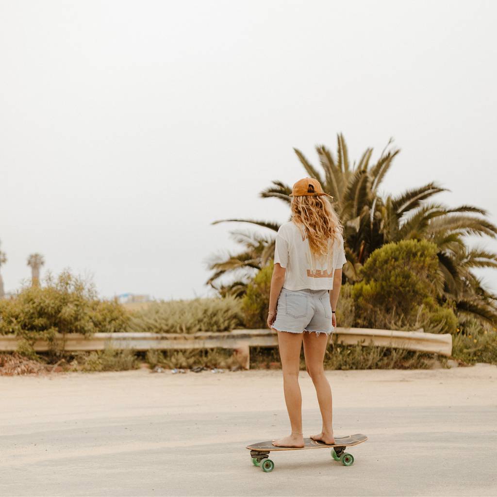 The image features a female model skateboarding wearing the Be Kind Vibes Desert Waves crop top and cut off jean shorts.