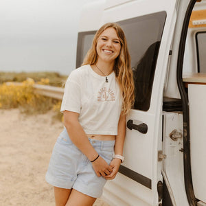 Hero image featuring a female model wearing the Be Kind Vibes Desert Waves crop top with jean shorts. The model is leaning against a white van smiling at the camera.