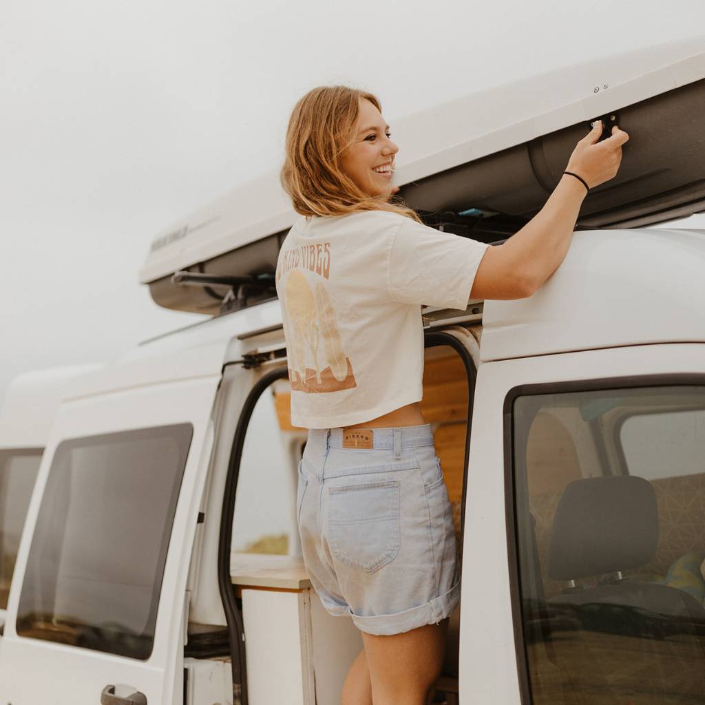 
                  
                    Image features a female model wearing the Be Kind Vibes Desert Waves crop top with jean shorts. The model is standing on the side of a van playing with the roof rack.
                  
                