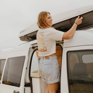 Image features a female model wearing the Be Kind Vibes Desert Waves crop top with jean shorts. The model is standing on the side of a van playing with the roof rack.
