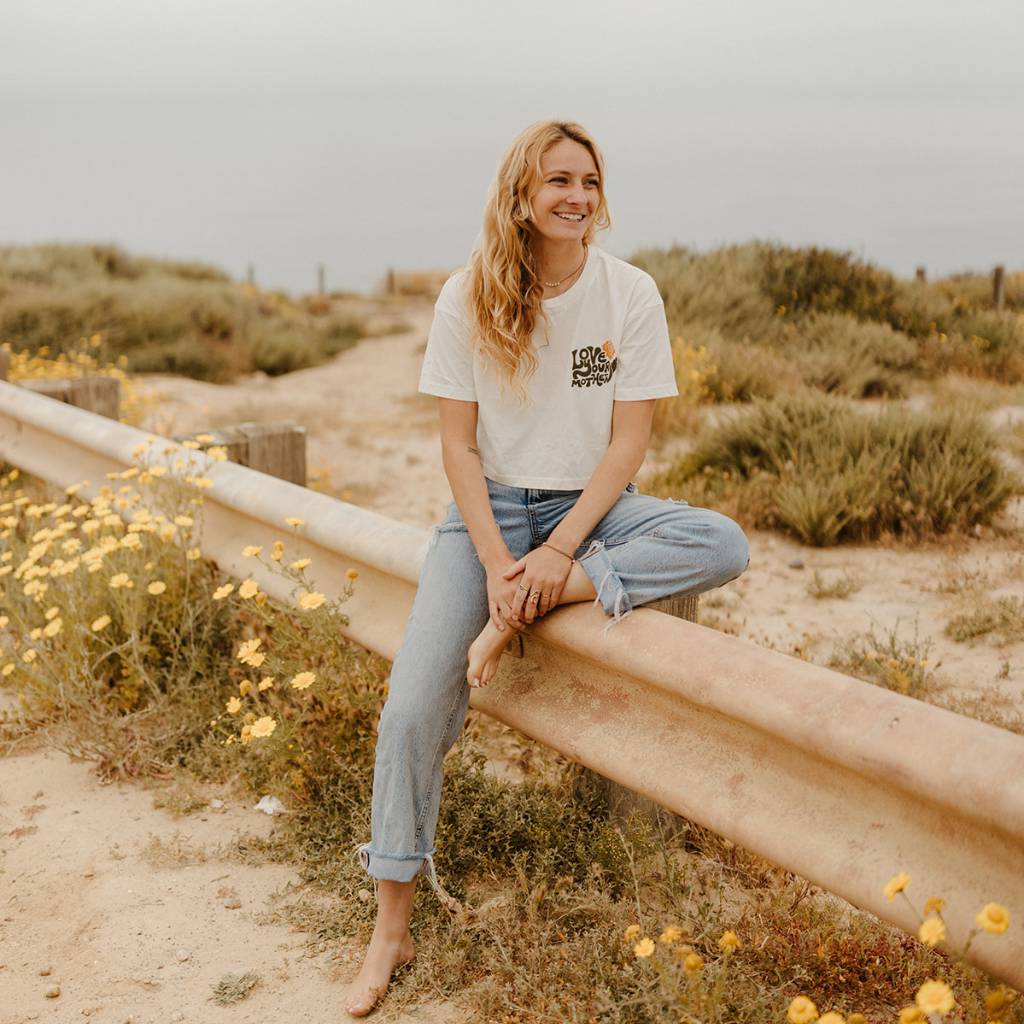 Hero image featuring a female model sitting on a guard rail wearing blue jeans and the Be Kind Vibes Love Your Mother crop top