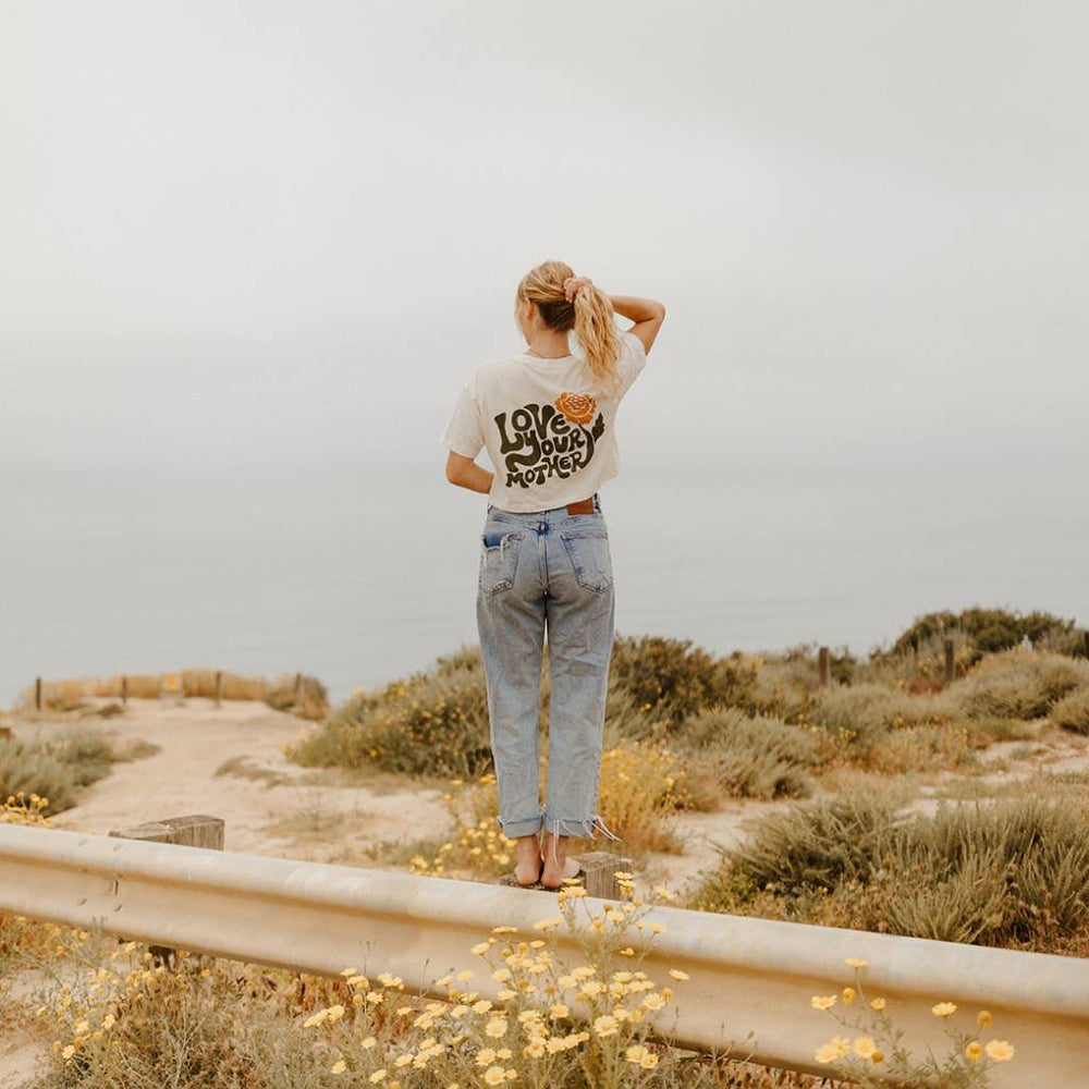 Image features a female model standing on a guard rail overlooking the ocean wearing blue jeans and the Be Kind Vibes Love Your Mother crop top.