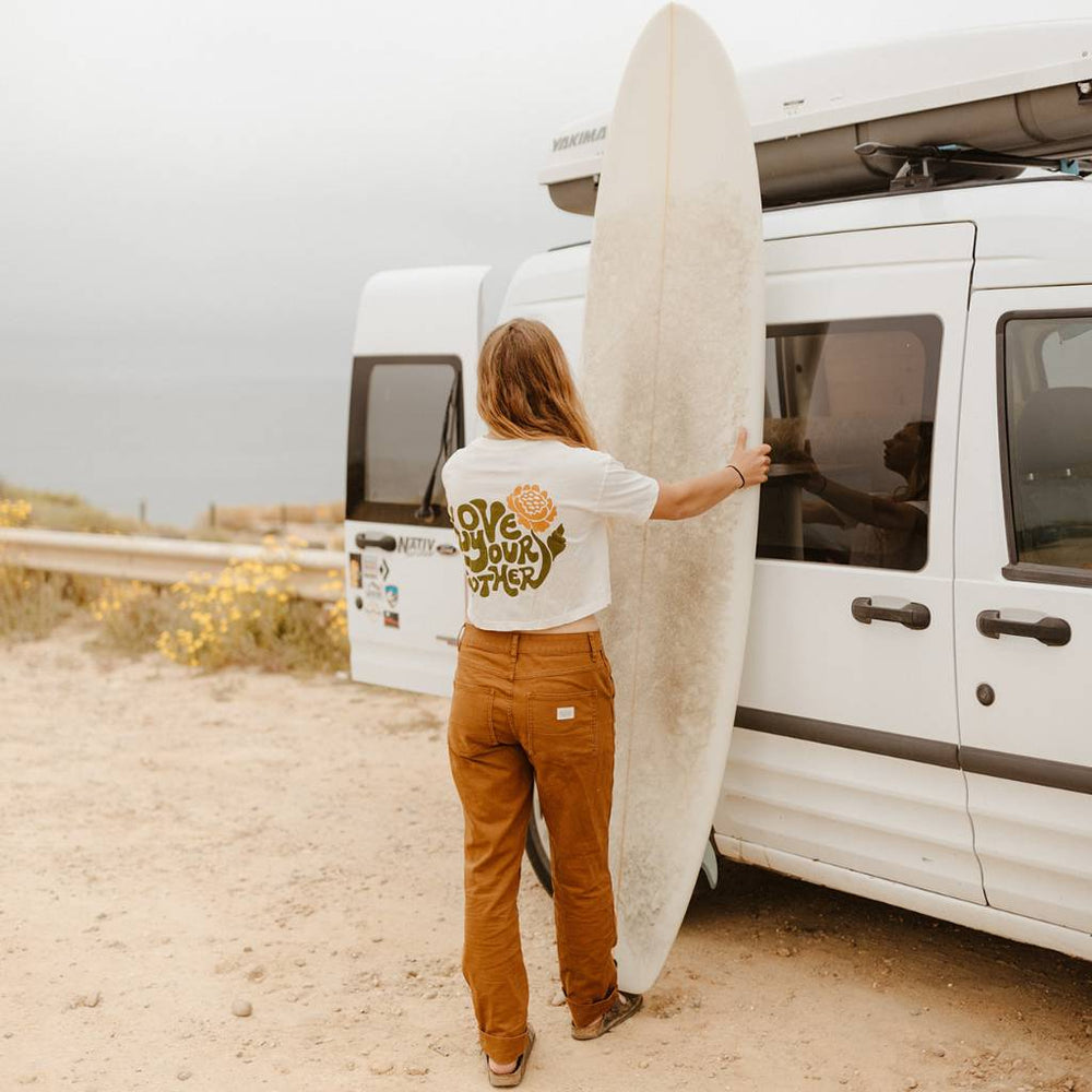 Image features a female model holding and looking at a white surfboard that's leaning up against a white van. The model is wearing the Be Kind Vibes Love Your Mother crop top with rust colored pants.