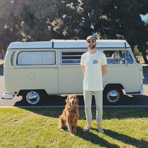 Image features the sustainably made 100% organic cotton Love Your Mother tee from Be Kind Vibes on a male model wearing sunglasses, a green hat, khaki pants and standing in the grass with a cute golden retriever in front of a VW bus.
