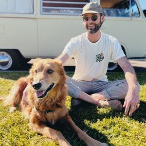 Image features the sustainably made 100% organic cotton Love Your Mother tee from Be Kind Vibes on a male model wearing sunglasses, a green hat, khaki pants and sitting in the grass with a cute golden retriever. 