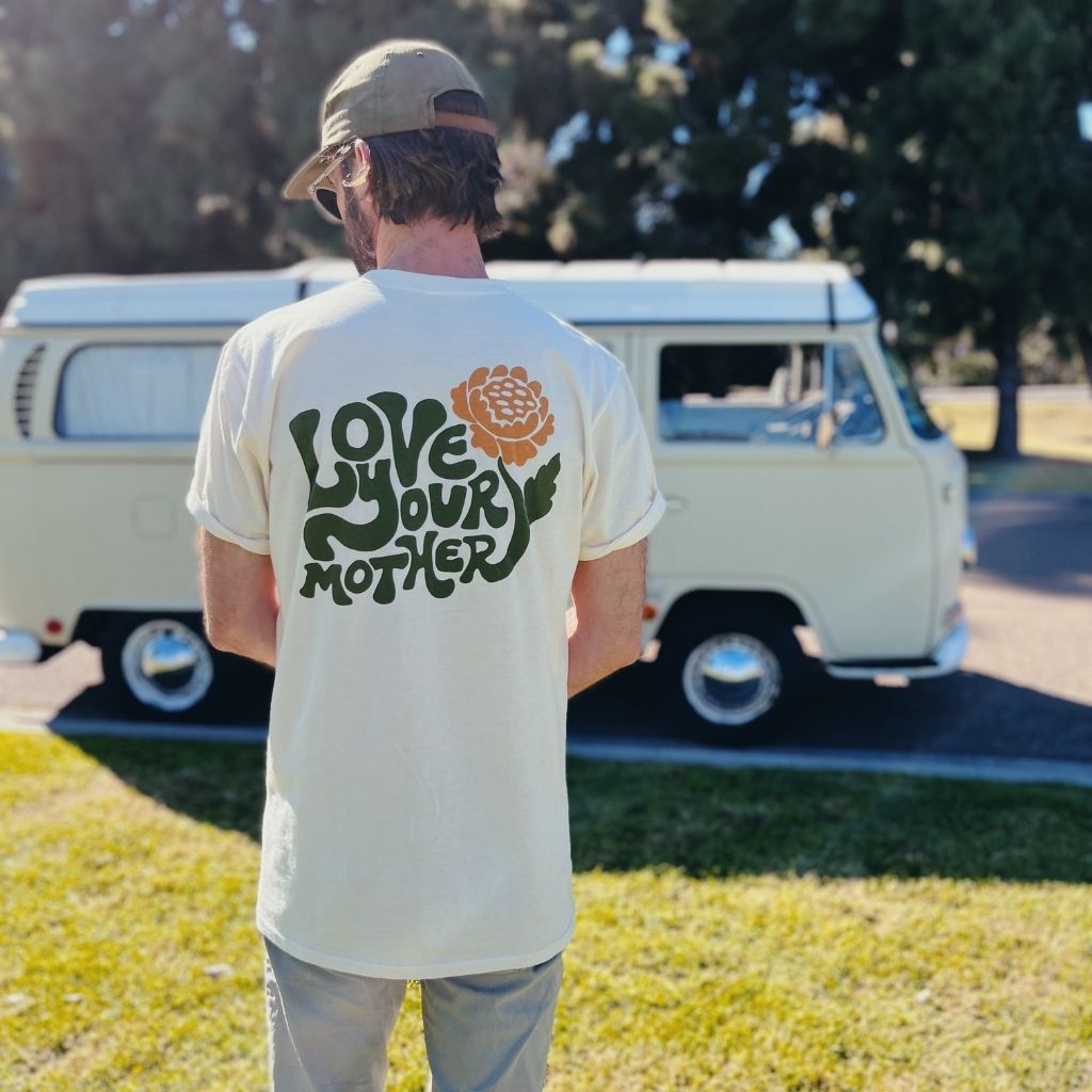 
                  
                    Hero image featuring the back of the 100% organic cotton Be Kind Vibes Love Your Mother tee in natural on a male model standing in front of a VW bus.
                  
                