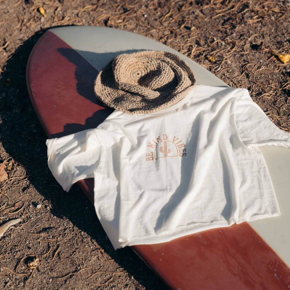 
                  
                    Image features the Be Kind Vibes Desert Waves crop top laying on top of a red and cream surfboard with a straw bucket hat.
                  
                