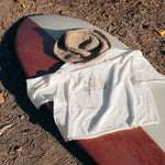 Image features the Be Kind Vibes Desert Waves crop top laying on top of a red and cream surfboard with a straw bucket hat.