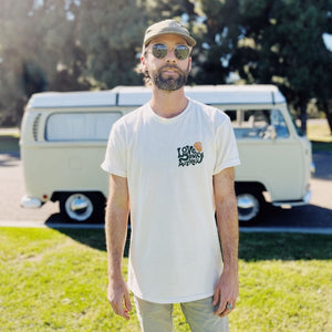 Hero image featuring the front of the Love Your Mother tee in natural on a male model wearing sunglasses, a green hat, khaki pants, and standing in front of a VW bus.