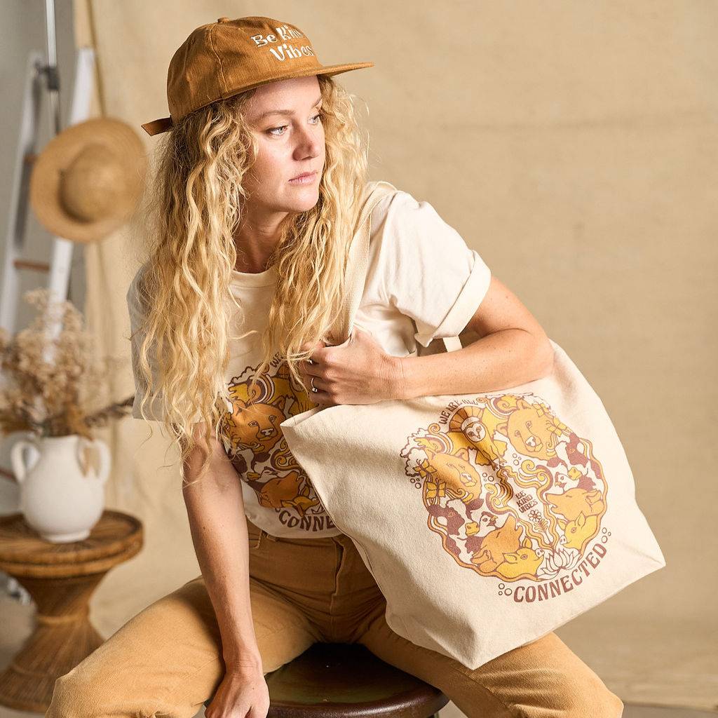 
                  
                    Image features a side angle photo of a female model sitting on a stool in front of a beige fabric backdrop. She's wearing a natural colored t-shirt, brown khaki pants, the Be Kind Hemp hat in rust, and is holding the Be Kind Vibes We Are All Connected tote bag over her shoulder.
                  
                