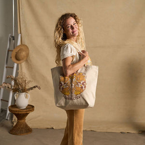 Image features a side angle photo of a female model standing in front of a beige fabric backdrop. She's wearing a natural colored t-shirt, brown khaki pants, and is holding the Be Kind Vibes We Are All Connected tote bag over her shoulder.