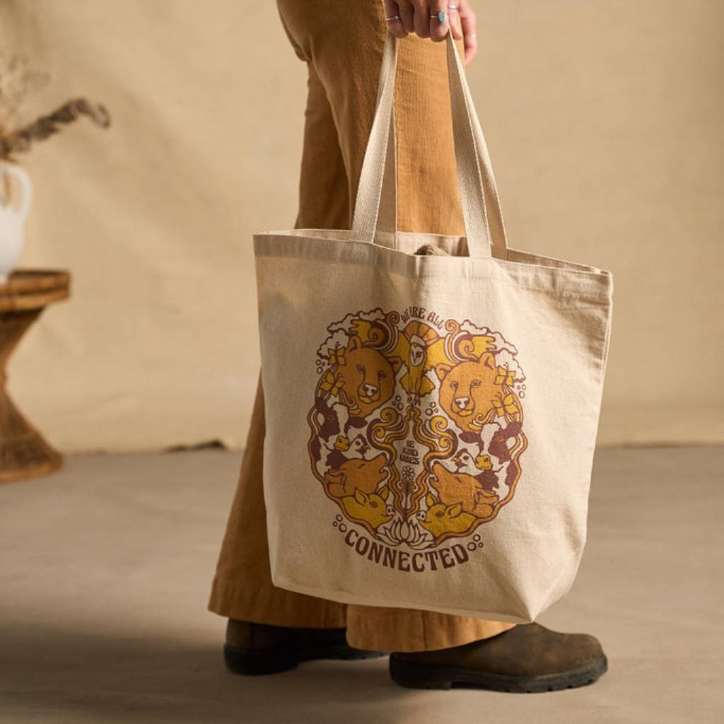 
                  
                    Image features a side angle shot of a female model wearing light brown khakis holding the Be Kind Vibes We Are All Connected tote bag in natural off to the side.
                  
                