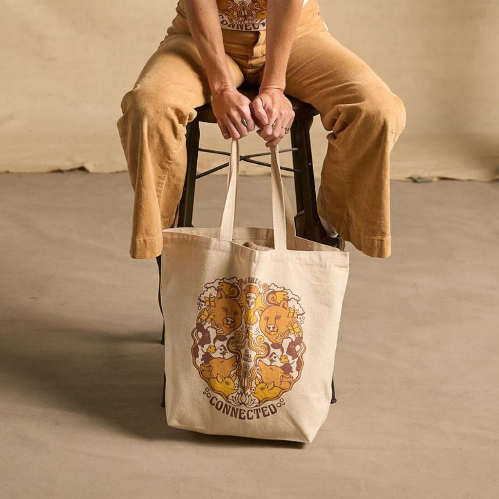 
                  
                    Hero image featuring a female model sitting on a stool wearing light brown khaki pants. She's holding the Be Kind Vibes We Are All Connected tote bag in natural in front of her.
                  
                