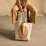 Hero image featuring a female model sitting on a stool wearing light brown khaki pants. She's holding the Be Kind Vibes We Are All Connected tote bag in natural in front of her.