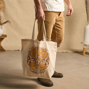 Hero image features a male model holding with one hand the Be Kind Vibes We Are All Connected tote bag. The design features wild animals and farm animals in various shades of earthy yellows and browns. The model is standing in front of a beige fabric backdrop with props.
