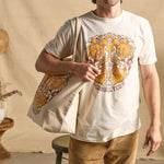 Image features a male model holding the Be Kind Vibes We Are All Connected tote bag over his right shoulder. He's wearing the We Are All Connected t-shirt, light brown khaki pants, and is standing in front of a beige fabric backdrop.