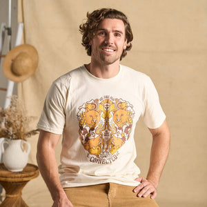 Image features a male model sitting on a stool in front of a beige fabric backdrop wearing the Be Kind Vibes We Are All Connected t-shirt which features an animal design on the front in earth tones including bears, an owl, pigs, cows, wolves, and chickens.