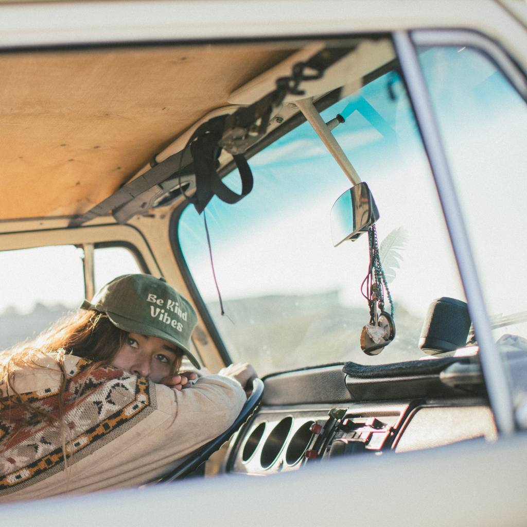
                  
                    Image features a female model leaning against the steering wheel of a VW bus wearing the cactus colored Be Kind Hemp Hat and a khaki jacket.
                  
                