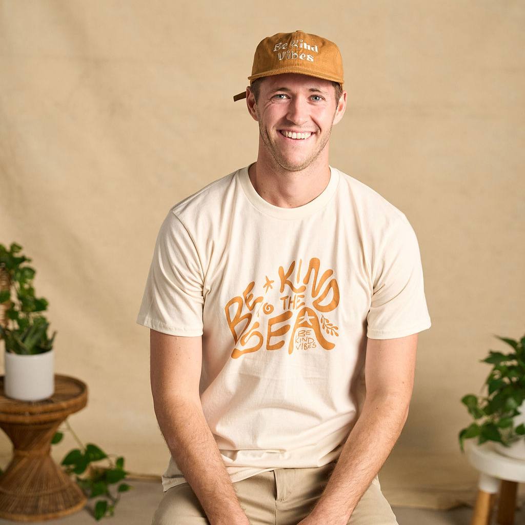 Image features a male model sitting on a stool with plants in the background wearing the rust colored Be Kind Vibes Be Kind Hemp hat, khaki pants, and the Be Kind Vibes Be Kind to the Sea organic cotton t-shirt in a natural colorway.