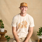 Image features a male model sitting on a stool with plants in the background wearing the rust colored Be Kind Vibes Be Kind Hemp hat, khaki pants, and the Be Kind Vibes Be Kind to the Sea organic cotton t-shirt in a natural colorway.