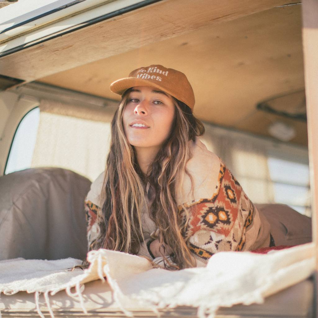 Image features a female model laying inside a VW bus wearing the rust colored Be Kind Vibes Be Kind Hemp Hat.