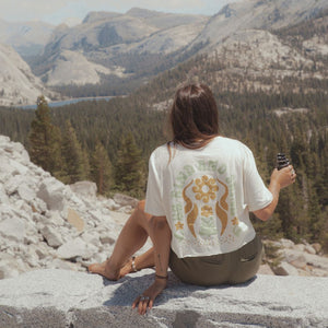 Image features a female model holding binoculars looking out over the Yosemite Valley wearing the sustainably made hemp and organic cotton crop top in natural by Be Kind Vibes. 