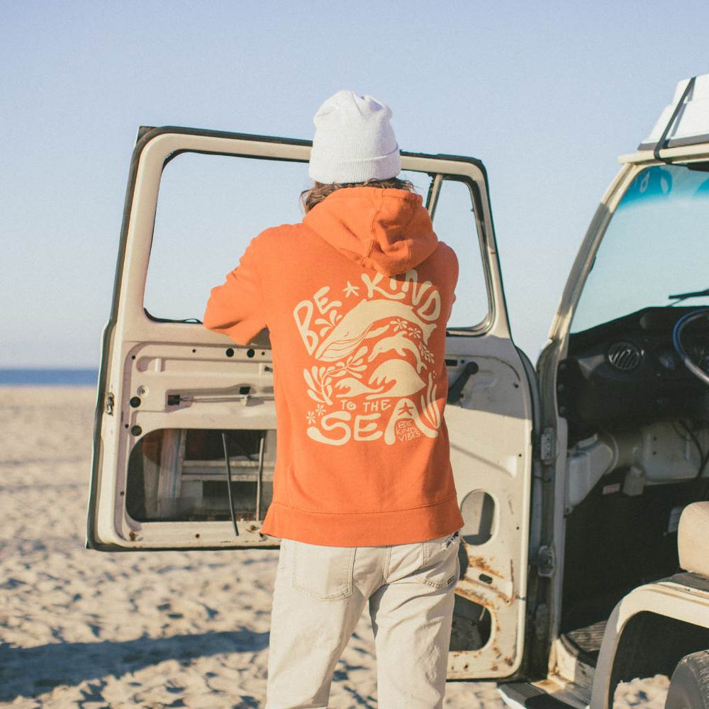 
                  
                    Image features  the back of a male model looking out over the ocean, standing next to a VW bus wearing the Be Kind Vibes 100% organic cotton hoodie in burnt orange. 
                  
                