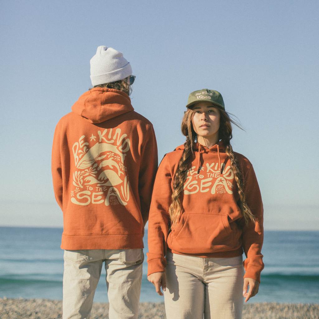 Image features the back of a male model and the front of a female model who are standing by the ocean wearing the Be Kind Vibes 100% organic cotton To the Sea Hoodie in burnt orange.