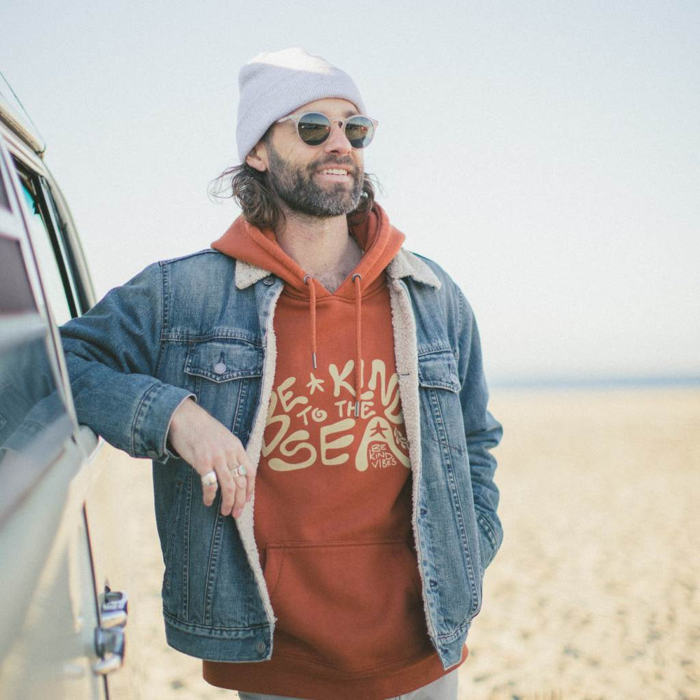 
                  
                    Image features a male model leaning on a VW bus wearing a grey beanie, a jean jacket, and the Be Kind Vibes 100% organic cotton To the Sea hoodie underneath.
                  
                