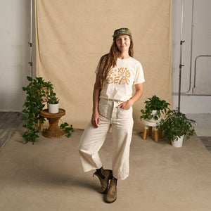 Image features a female model  wearing the Be Kind Vibes organic cotton To the Sea t-shirt with white pants and brown boots. In the background is a natural colored drop cloth with various sized plants.