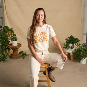 Image features a female model sitting on a wooden stool wearing the Be Kind Vibes organic cotton To the Sea t-shirt with white pants and brown boots. In the background is a natural colored drop cloth with various sized plants.