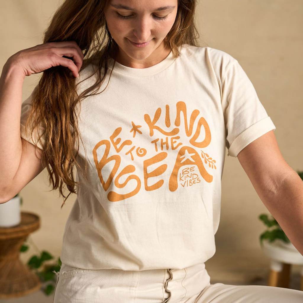 Image features a female model sitting on a wooden stool wearing the Be Kind Vibes organic cotton To the Sea t-shirt with white pants and brown boots. In the background is a natural colored drop cloth with various sized plants.