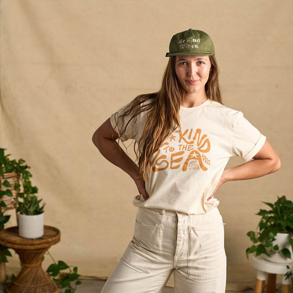 Image features a female model  wearing the Be Kind Vibes organic cotton To the Sea t-shirt with white pants and brown boots. In the background is a natural colored drop cloth with various sized plants.