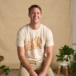 Image features a male model  wearing the Be Kind Vibes organic cotton To the Sea t-shirt with khaki pants. In the background is a natural colored drop cloth with various sized plants.
