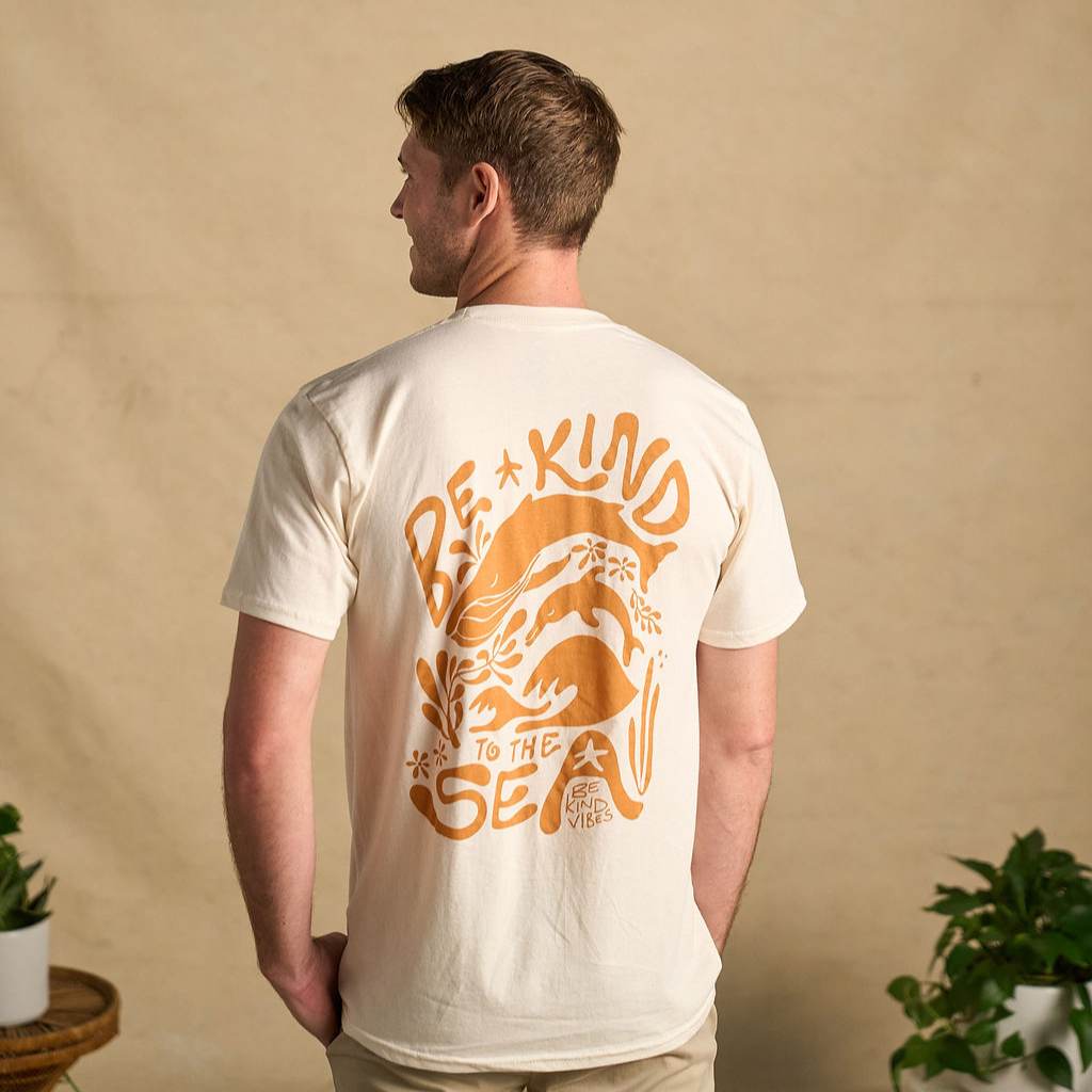 Image features the back of a male model  wearing the Be Kind Vibes organic cotton To the Sea t-shirt with khaki pants. In the background is a natural colored drop cloth with various sized plants.