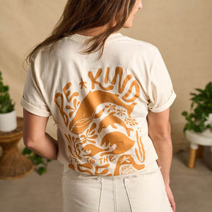 Image features the back of a female model  wearing the Be Kind Vibes organic cotton To the Sea t-shirt with white pants and brown boots. In the background is a natural colored drop cloth with various sized plants.