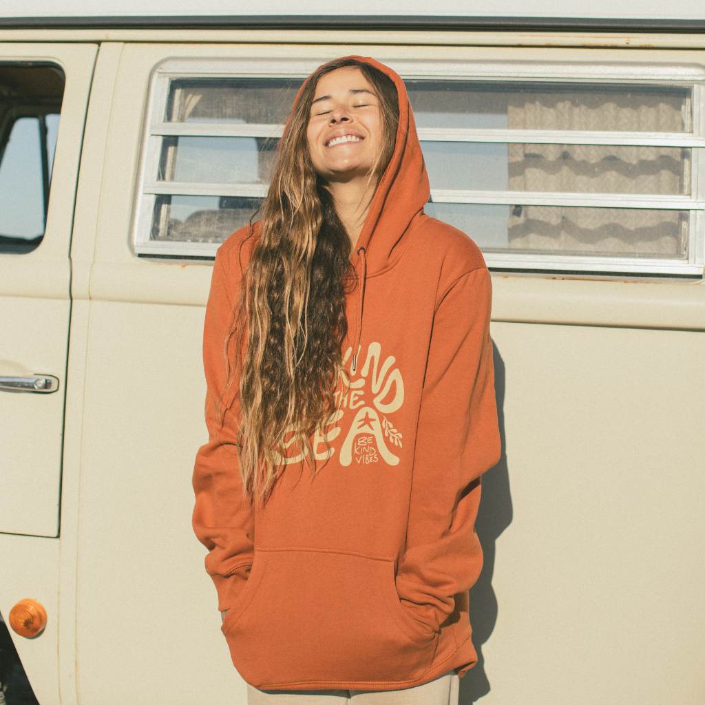 Image features a female model standing in front of a VW Bus wearing the Be Kind Vibes To the Sea hoodie in burnt orange with khaki pants.