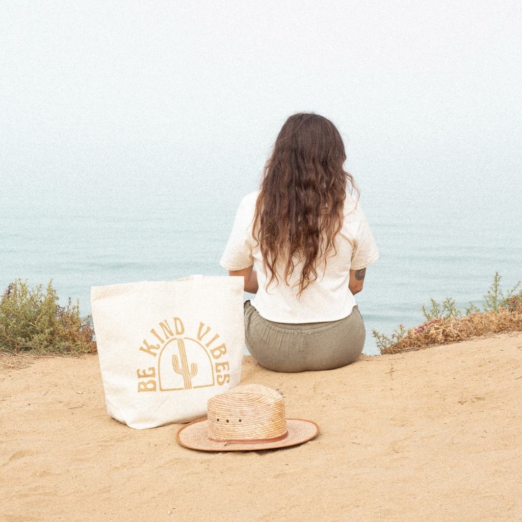Image featuring a female model sitting on a cliff over looking the ocean with the Be Kind Vibes Desert Tote bag resting behind her next to a straw hat.