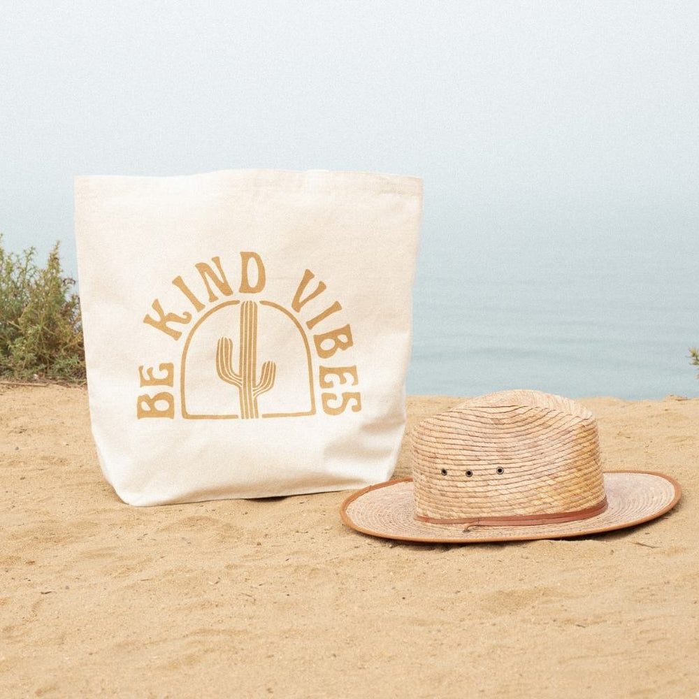 
                  
                    Image featuring the Be Kind Vibes sustainable Desert Tote sitting in the sand next to a straw hat overlooking the ocean.
                  
                