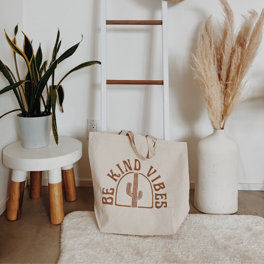 
                  
                    Hero image featuring the Be Kind Vibes ethically made Desert Tote resting on the ground surrounded by plants and a white rug.
                  
                