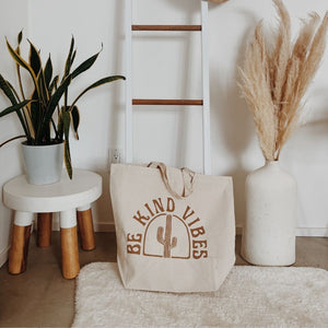 Hero image featuring the Be Kind Vibes ethically made Desert Tote resting on the ground surrounded by plants and a white rug.