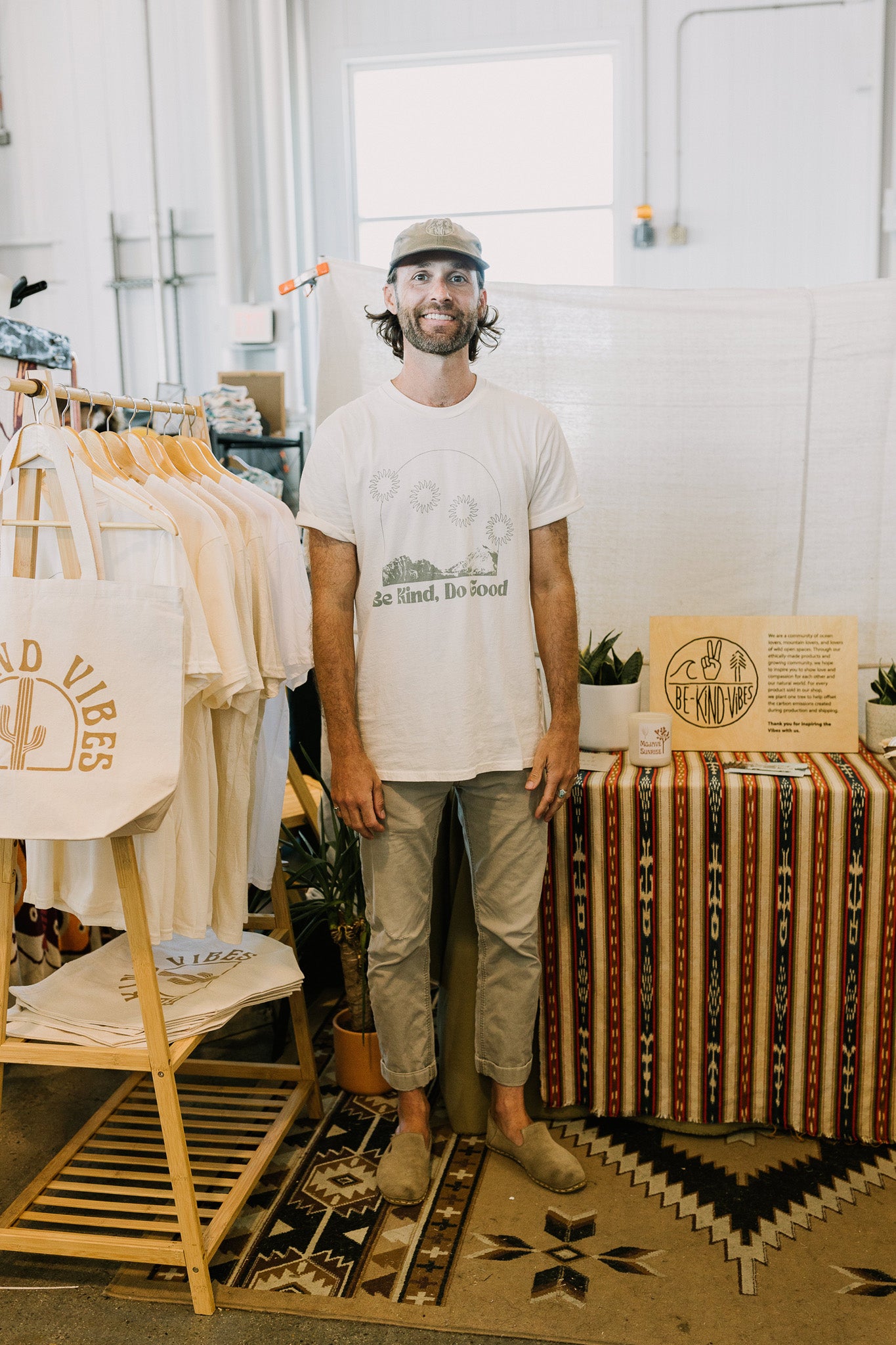 Be Kind Vibes Owner and founder standing in front of a Be Kind Vibes pop-up shop wearing a white Be Kind, Do Good t-shirt, khaki pants, and a green hat.